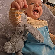 Elio is registered to the contest to win money with this photo: baby, child, smiling, stuffed_animal, toy, blanket, blue_blanket, plush, indoor, happy, face, hand, clothing, overalls, striped_sleeves, elephant_pattern, furniture, couch, person, portrait