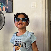 Ousmane a rejoint le concours — aidez-le/la à gagner de superbes lots ! child, smiling, sunglasses, braided_hair, tshirt, teddy_bear_print, indoor, wall, fan, picture_frame, happy, person, portrait, casual_clothing, standing, short_sleeve, light_blue, decor, room, young_child