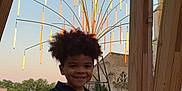 Ousmane a rejoint le concours — aidez-le/la à gagner de superbes lots ! child, smiling, curly_hair, black_clothing, juice_pouch, wooden_bench, outdoor, grass, light_installation, art, building, stone_wall, trees, evening, playful, portrait, happy, person, nature, relaxing