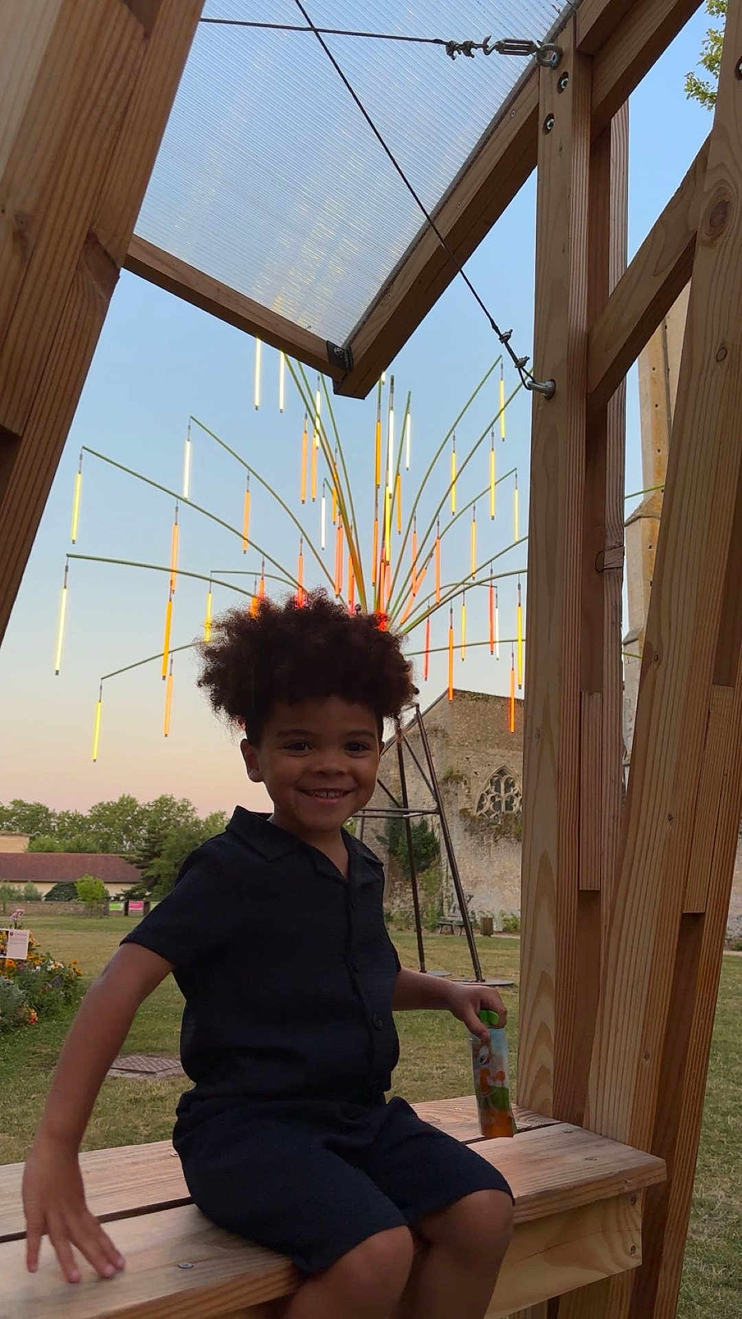 Ousmane a rejoint le concours — aidez-le/la à gagner de superbes lots ! child, smiling, curly_hair, black_clothing, juice_pouch, wooden_bench, outdoor, grass, light_installation, art, building, stone_wall, trees, evening, playful, portrait, happy, person, nature, relaxing