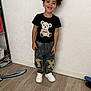 Ousmane a rejoint le concours — aidez-le/la à gagner de superbes lots ! child, toddler, smiling, standing, indoor, curly_hair, black_tshirt, teddy_bear, denim_jeans, patched_clothing, white_shoes, wooden_floor, wall, toy, shelf, happy, person, casual_clothing, floor, playful