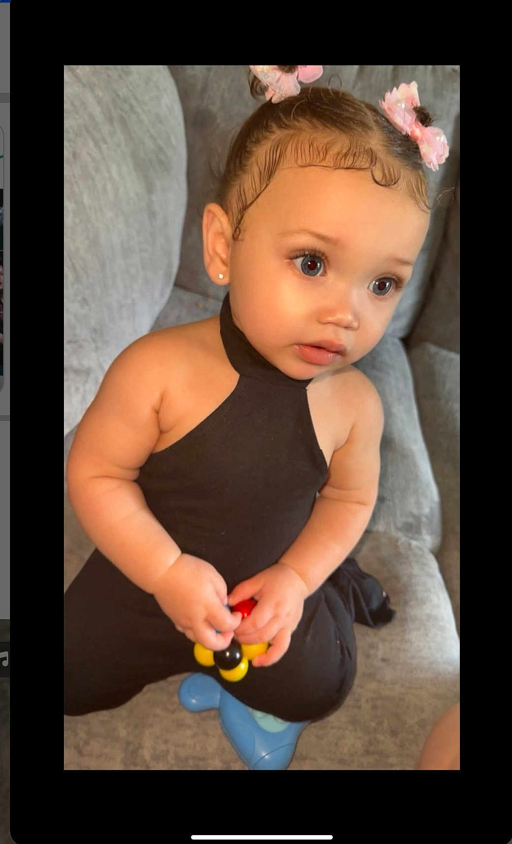 Amora Lei' is registered to the contest to win money with this photo: toddler, child, baby, black_outfit, pink_bows, toy, couch, gray_furniture, curly_hair, baby_hairs, earrings, indoor, portrait, cute, sitting, small_hands, expression, looking_away, soft_lighting, casual