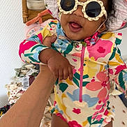 Ely a rejoint le concours — aidez-le/la à gagner de superbes lots ! baby, child, sunglasses, hat, colorful_clothing, floral_pattern, hand, indoor, smiling, happy, skin, person, infant, baby_swimsuit, wicker_shelf, bottles, blanket, foot, face, cute