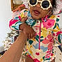 baby, child, sunglasses, hat, colorful_clothing, floral_pattern, hand, indoor, smiling, happy, skin, person, infant, baby_swimsuit, wicker_shelf, bottles, blanket, foot, face, cute