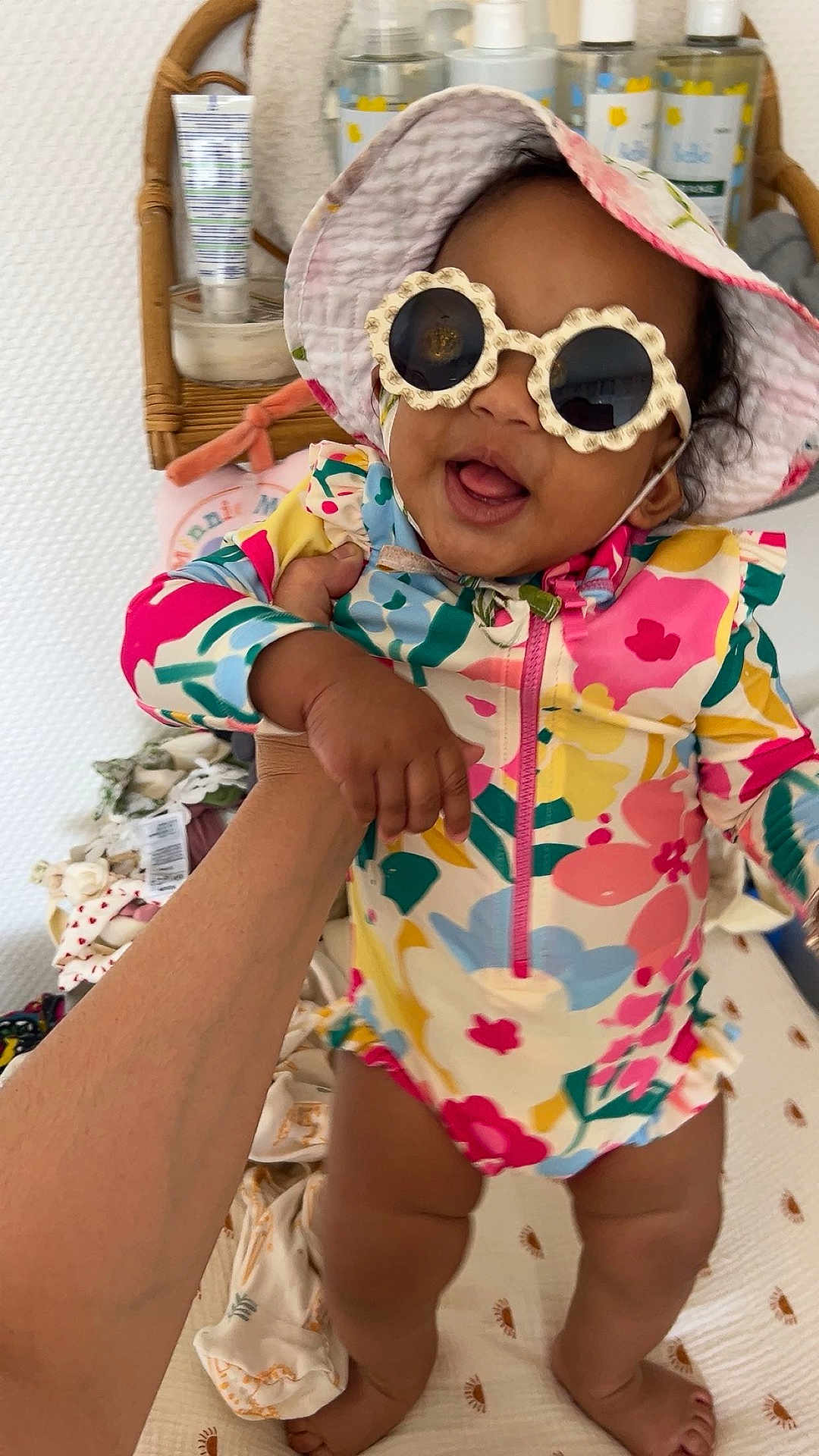 Ely a rejoint le concours — aidez-le/la à gagner de superbes lots ! baby, child, sunglasses, hat, colorful_clothing, floral_pattern, hand, indoor, smiling, happy, skin, person, infant, baby_swimsuit, wicker_shelf, bottles, blanket, foot, face, cute