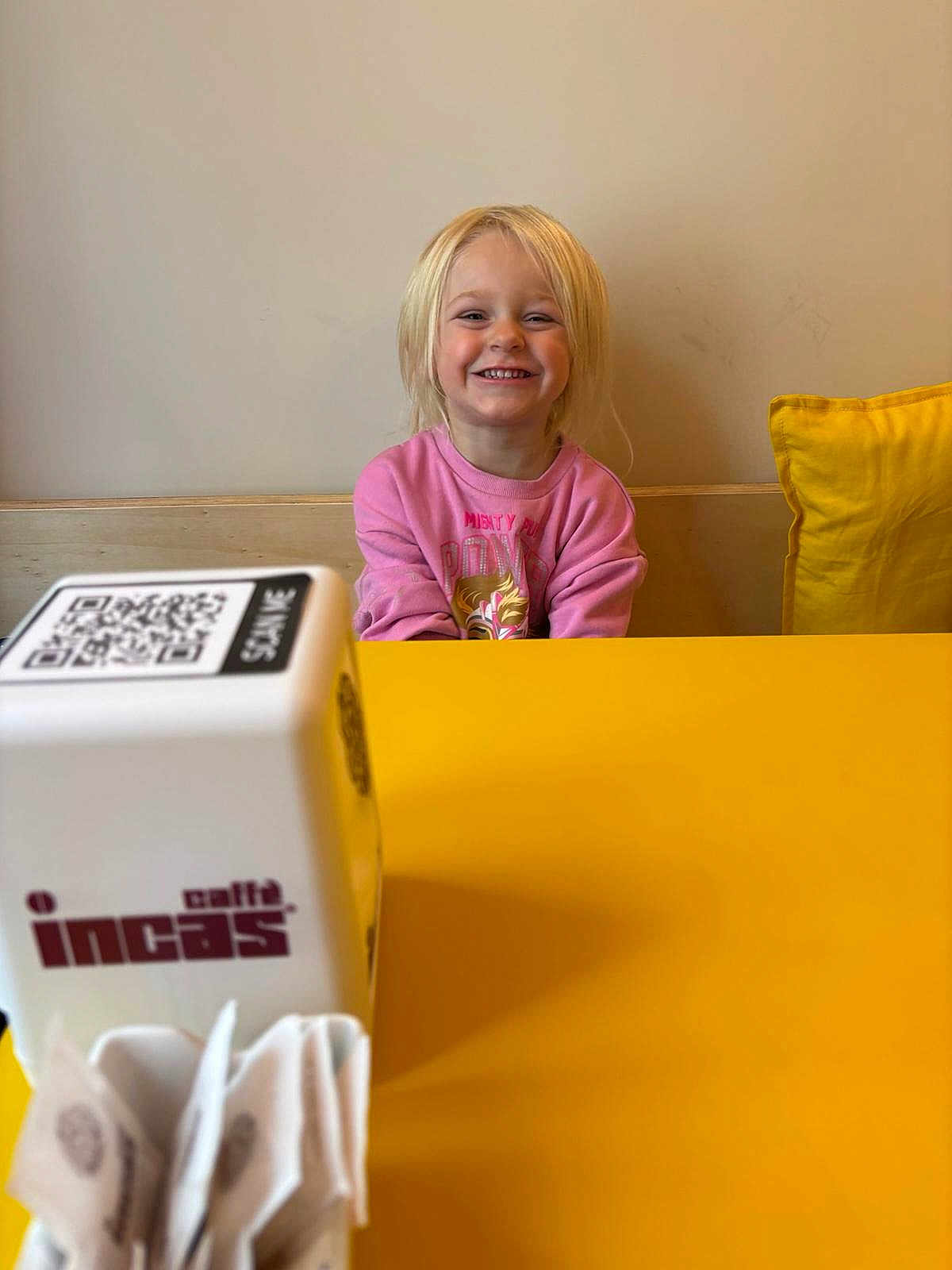 Inaya a rejoint le concours — aidez-le/la à gagner de superbes lots ! child, smiling, blonde_hair, pink_sweater, yellow_table, cafe, qr_code, white_cube, napkins, happy, indoor, person, table, wall, pillow, seat, casual, portrait, young, cute