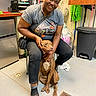 woman, dog, indoor, floor, trash_bin, sitting, smiling, sunglasses, casual_clothing, shelf, bag, drain, tile_floor, short_sleeve_shirt, pants, shoe, pet, person, happy, companion