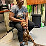 woman, dog, indoor, smiling, seated, casual_clothing, short_sleeves, eyewear, chair, floor, trash_bin, shelf, storage_room, happy, pet, brown_dog, black_pants, sneakers, leash, relaxed