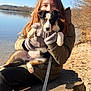 puppy, person, gloves, jacket, lake, water, tree_stump, outdoor, sunlight, dog, animal, nature, fall, leash, sitting, brown_hair, calm, sky, bare_trees, young_person