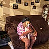 person, dog, beagle, couch, brown_cover, indoor, wooden_floor, family_photos, picture_frames, slippers, tie_dye_shirt, hugging, pet, home_decor, wall_art, smiling_dog, living_room, cozy, tongue_out, seated