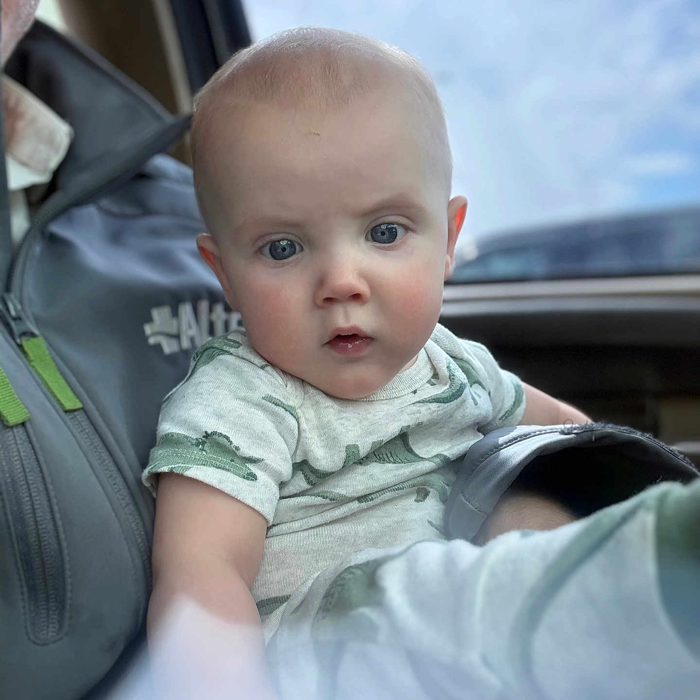 Noah joined the competition — help win amazing prizes! baby, infant, child, person, face, blue_eyes, short_hair, clothing, shirt, car_interior, window, sky, clouds, adult_partial, lap, hand, expression, portrait, seat, casual