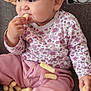 baby, child, snack, food, pink_pants, floral_shirt, sitting, couch, blanket, cartoon, munching, hand, face, cute, indoor, playful, snack_food, infant, clothing, happy