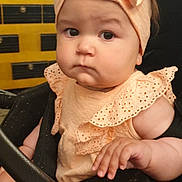 Klaudia is registered to the contest to win money with this photo: baby, child, stroller, headband, bow, peach_color, frilly_clothing, hand, face, indoor, infant, cute, serious_expression, person, young_child, sitting, closeup, portrait, skin, furniture