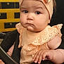 baby, child, stroller, headband, bow, peach_color, frilly_clothing, hand, face, indoor, infant, cute, serious_expression, person, young_child, sitting, closeup, portrait, skin, furniture