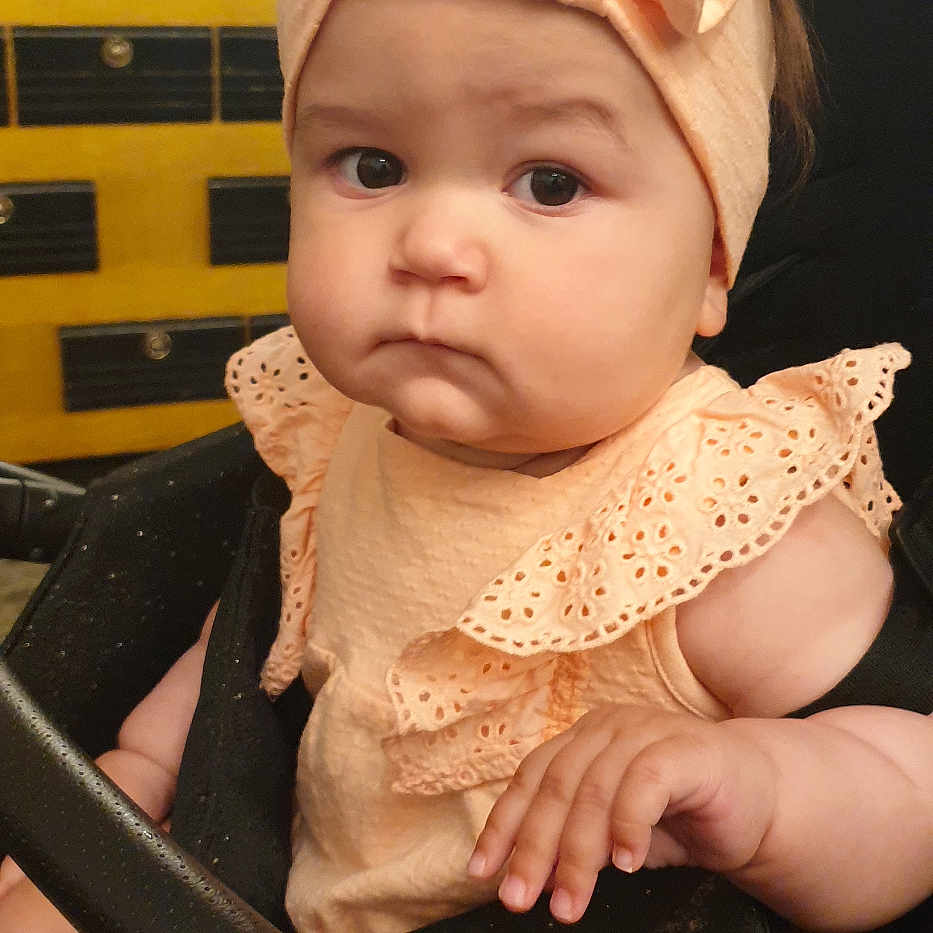 Klaudia is registered to the contest to win money with this photo: baby, bow, child, closeup, cute, face, frilly_clothing, furniture, hand, headband, indoor, infant, peach_color, person, portrait, serious_expression, sitting, skin, stroller, young_child