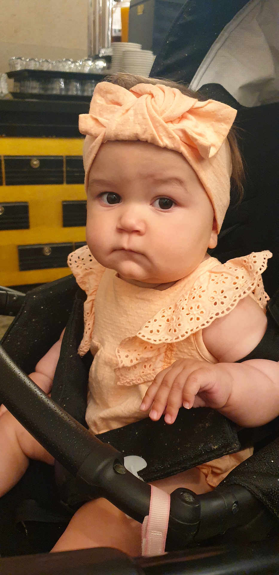 Klaudia is registered to the contest to win money with this photo: baby, child, stroller, headband, bow, peach_color, frilly_clothing, hand, face, indoor, infant, cute, serious_expression, person, young_child, sitting, closeup, portrait, skin, furniture