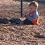 Ryker is registered to the contest to win money with this photo: child, toddler, playground, wood_chips, toy_car, sitting, side_profile, orange_shirt, long_sleeve, sunlight, outdoor, grass, concrete_wall, metal_pole, playtime, wheels, casual_clothing, candid, kid_playing, nature