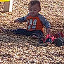 child, toddler, boy, playground, wood_chips, mulch, toy_car, orange_shirt, long_sleeves, sitting, bare_feet, face, hair, outdoor, sunlight, shadow, toy, playing, ground, casual_clothing