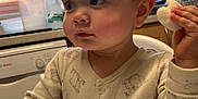 Finley is registered to the contest to win money with this photo: baby, child, high_chair, kitchen, pajamas, snack, food, face, hand, window, light, curious, cute, indoor, table, milk_jug, dishwasher, cabinet, person, expression