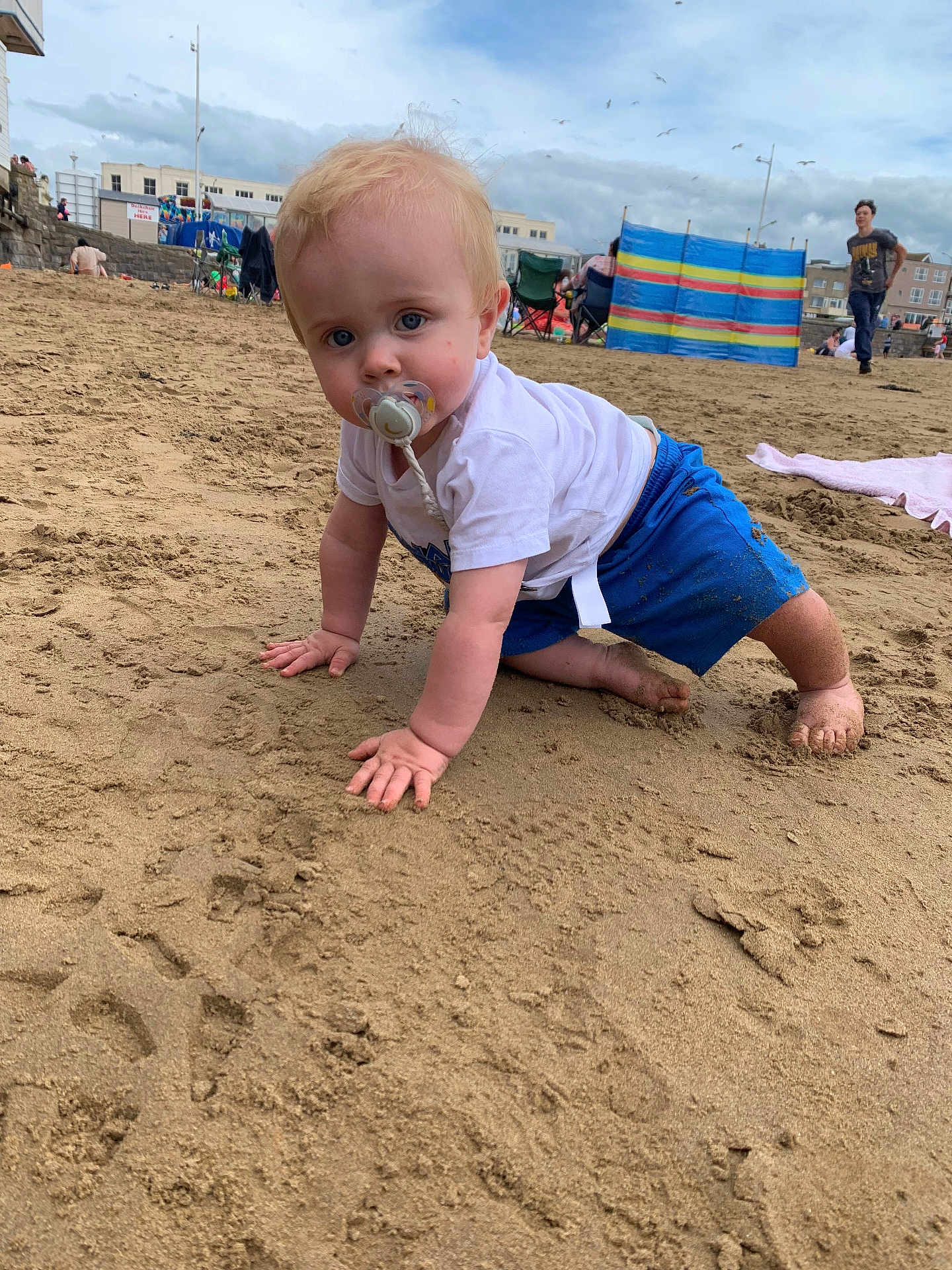 Finley joined the competition — help win amazing prizes! baby, child, pacifier, beach, sand, blue_shorts, white_tshirt, crawling, outdoor, cloudy_sky, people, windbreak, barefoot, playful, summer, vacation, person, footprints, toddler, casual
