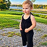 child, toddler, portrait, outdoor, standing, smiling, black_outfit, white_shoes, gravel, grass, lake, mist, hills, landscape, blue_sky, sunlight, trees, nature, fashion, earrings