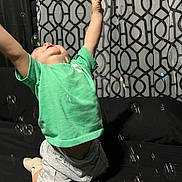 Amir joined the competition — help win amazing prizes! toddler, child, green_shirt, shorts, kneeling, bubbles, indoor, curtain, patterned_curtain, black_background, happy, arms_raised, playful, smiling, light_gray, socks, candid, joyful, person, home