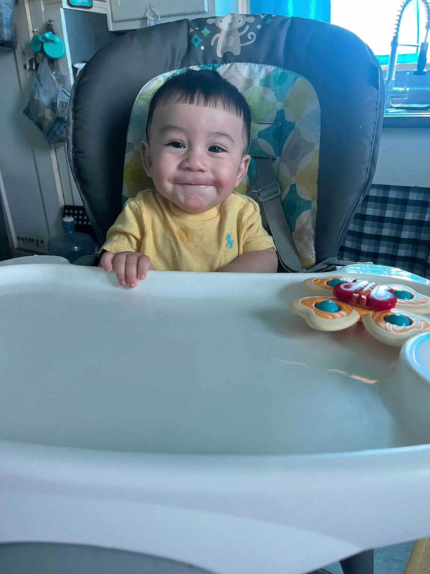 Amir is registered to the contest to win money with this photo: baby, high_chair, smile, yellow_shirt, toy, kitchen, child, face, expression, indoor, seat, feeding, person, hair, furniture, cute, happy, table, playful, infant