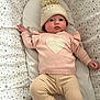 baby, infant, hat, knit_hat, pom_pom, pink_sweater, heart_pattern, star_pattern, blanket, lying_down, cute, child, soft_lighting, cozy, warm_clothing, portrait, indoors, face, expression, comfortable