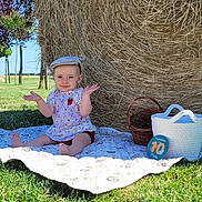 Maëlia a rejoint le concours — aidez-le/la à gagner de superbes lots ! baby, child, hat, blanket, grass, hay_bale, basket, outdoor, sunny, smile, dress, blue_sky, nature, summer, field, sitting, person, greenery, playful, cute