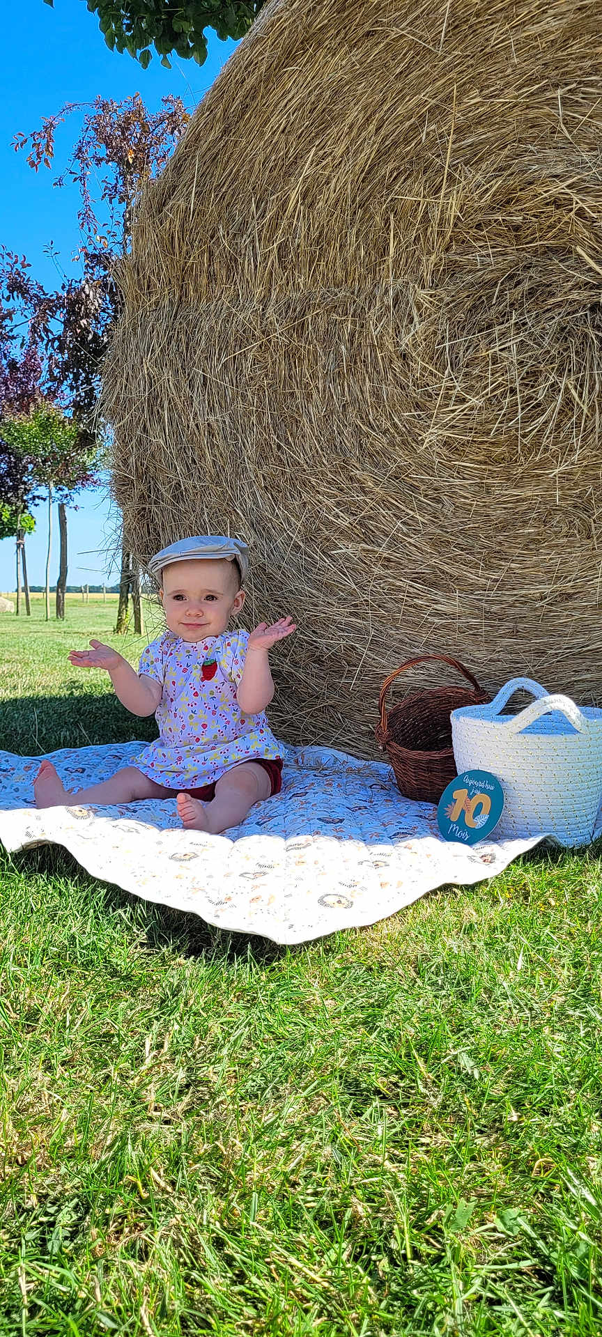 Maëlia a rejoint le concours — aidez-le/la à gagner de superbes lots ! baby, child, hat, blanket, grass, hay_bale, basket, outdoor, sunny, smile, dress, blue_sky, nature, summer, field, sitting, person, greenery, playful, cute