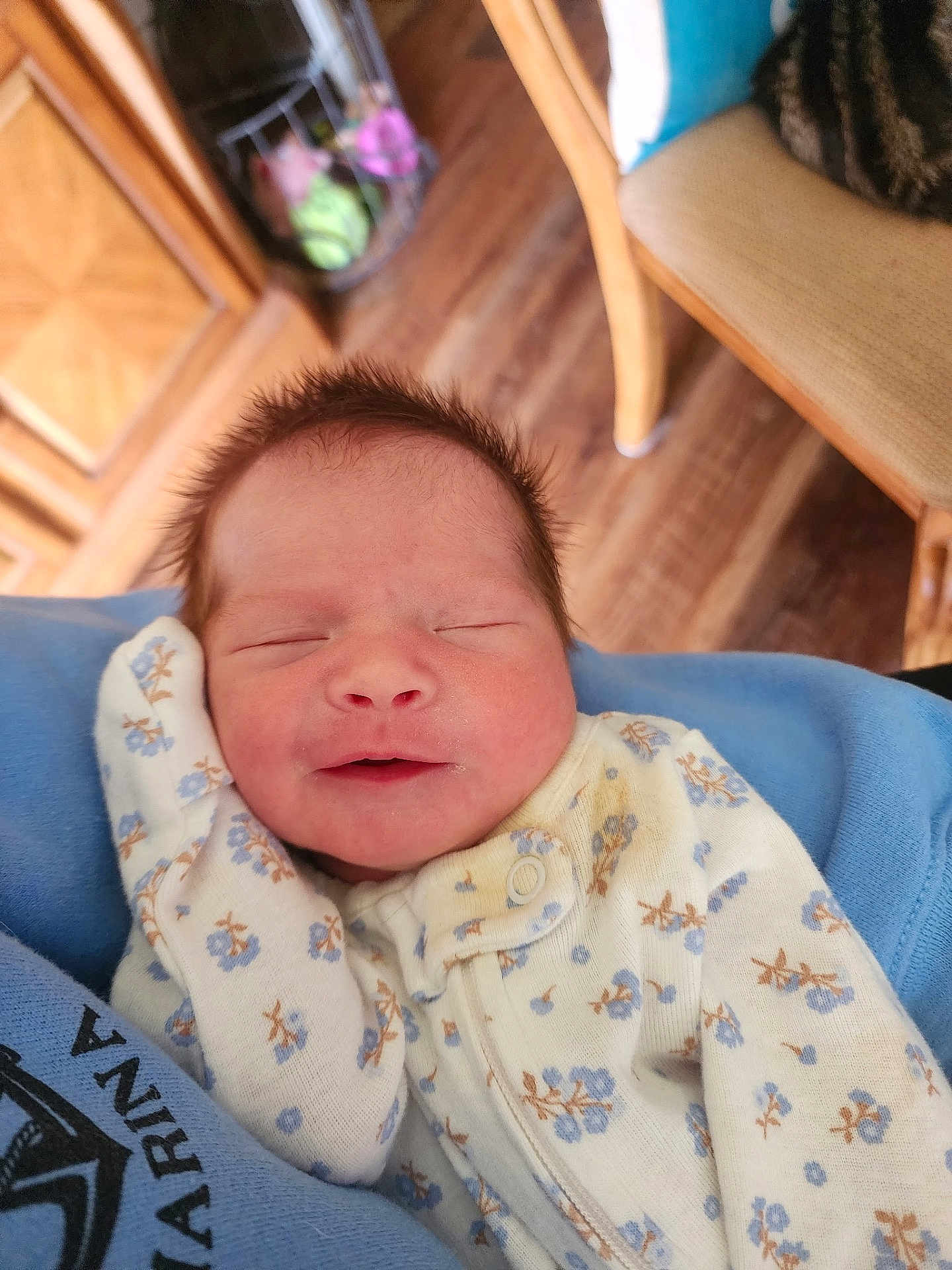 Cora is registered to the contest to win money with this photo: baby, newborn, infant, sleeping, smiling, floral_onesie, blue_blanket, hand, face, closed_eyes, soft_hair, cradled, lap, wooden_floor, chair, furniture, indoor, portrait, cozy, caregiver
