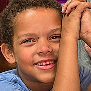 Damian is registered to the contest to win money with this photo: arm, blue_shirt, books, boxes, casual, child, close_up, curly_hair, eyes, face, happy, home, indoor, portrait, raised_arm, shelves, sitting, smiling, teeth, wood