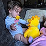 child, toddler, baby, sitting, couch, plush_toy, dinosaur, yellow_toy, toy, indoors, living_room, blanket, portrait, face, profile, brown_hair, long_sleeves, playtime, family, cute