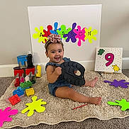 Alisson is registered to the contest to win money with this photo: baby, child, smiling, headband, toy_blocks, paint_cans, colorful, carpet, sitting, indoor, canvas, number_nine, easel, playtime, celebration, flower, happy, room, floor, person