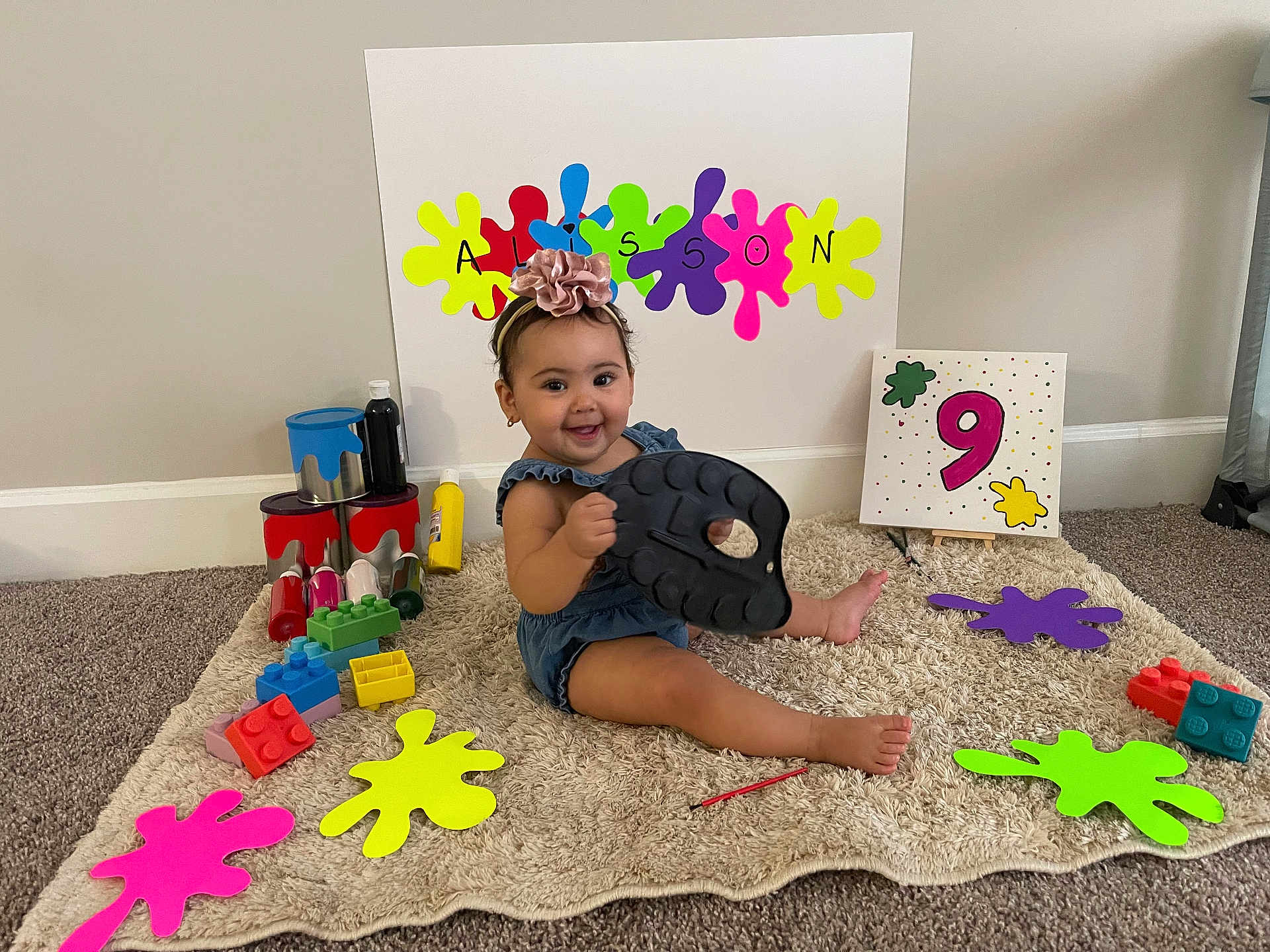 Alisson is registered to the contest to win money with this photo: baby, child, smiling, headband, toy_blocks, paint_cans, colorful, carpet, sitting, indoor, canvas, number_nine, easel, playtime, celebration, flower, happy, room, floor, person