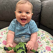 Walker is registered to the contest to win money with this photo: baby, smiling, toy, action_figure, couch, cushion, indoor, child, happy, shirt, blue_shirt, hand, face, person, sitting, playful, infant, young_child, home, cute
