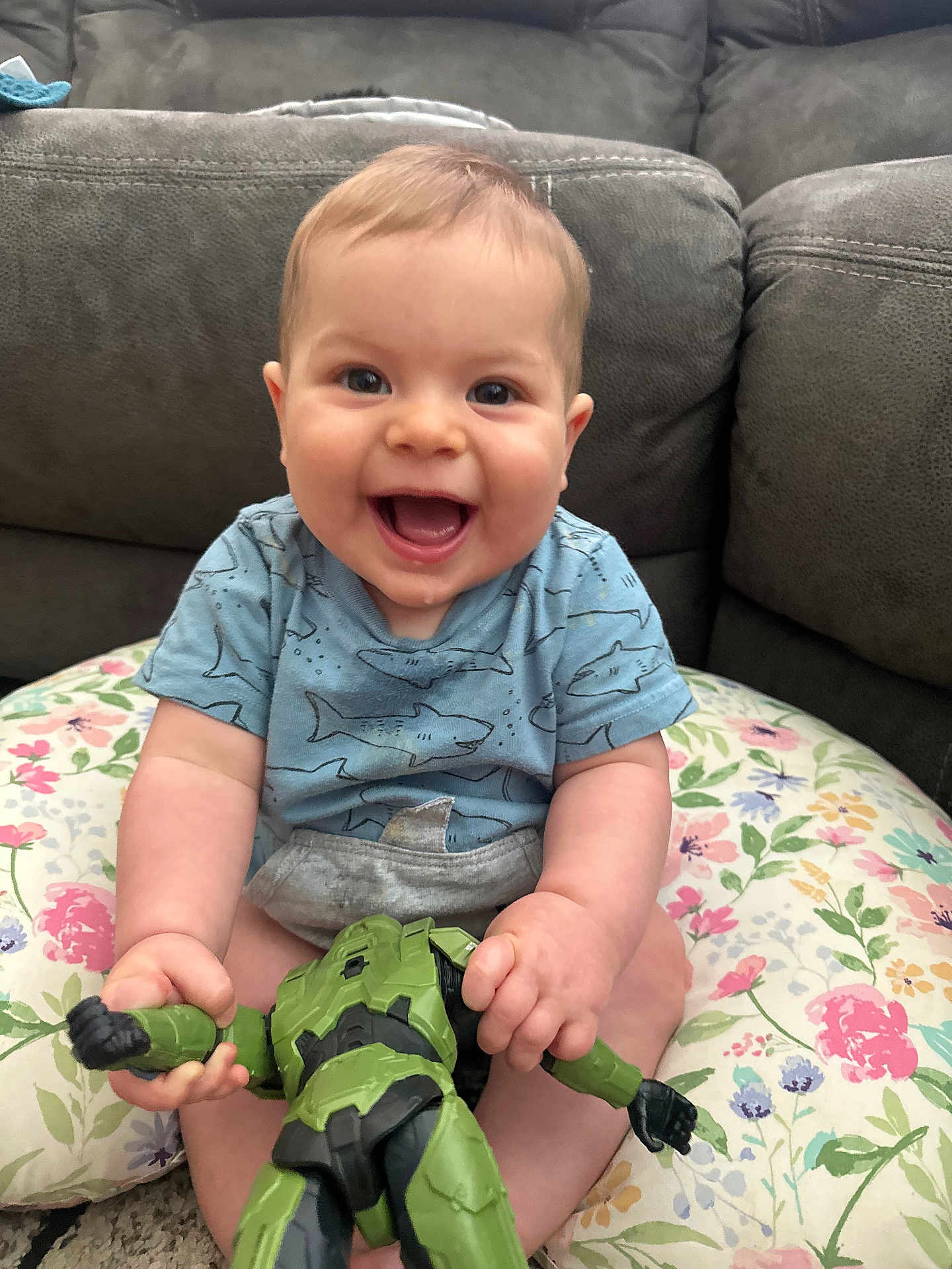 Walker is registered to the contest to win money with this photo: baby, smiling, toy, action_figure, couch, cushion, indoor, child, happy, shirt, blue_shirt, hand, face, person, sitting, playful, infant, young_child, home, cute