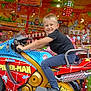 child, boy, smiling, motorbike, spider_man, amusement_ride, colorful, lights, fun, entertainment, toy, helmet_absent, casual_clothing, blue_pants, black_shirt, sneakers, indoor, background_characters, playful, happy