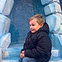 child, boy, puffer_jacket, blue_steps, stone_arch, sneakers, jeans, crouching, smiling, outdoor, playful, winter_clothing, frozen_theme, archway, casual, young, happy, portrait, fun, adventure