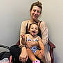 woman, child, toddler, laughing, smiling, indoor, armchair, casual_clothing, butterfly_swimsuit, pink_shoes, wooden_chair, happy, family, mother_and_child, affection, candid, joy, hair_bun, bracelet, skin_tone