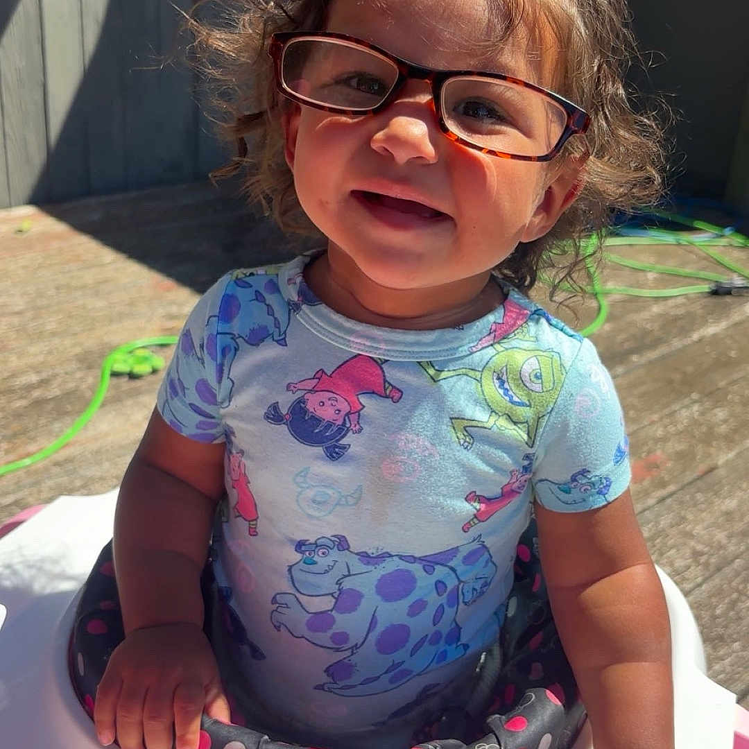 Kyelynn is registered to the contest to win money with this photo: baby, baby_walker, cartoon_print, child, curly_hair, cute, deck, flower_toys, glasses, hand, happy, outdoor, person, pink, play, shirt, smiling, summer, sunlight, toddler