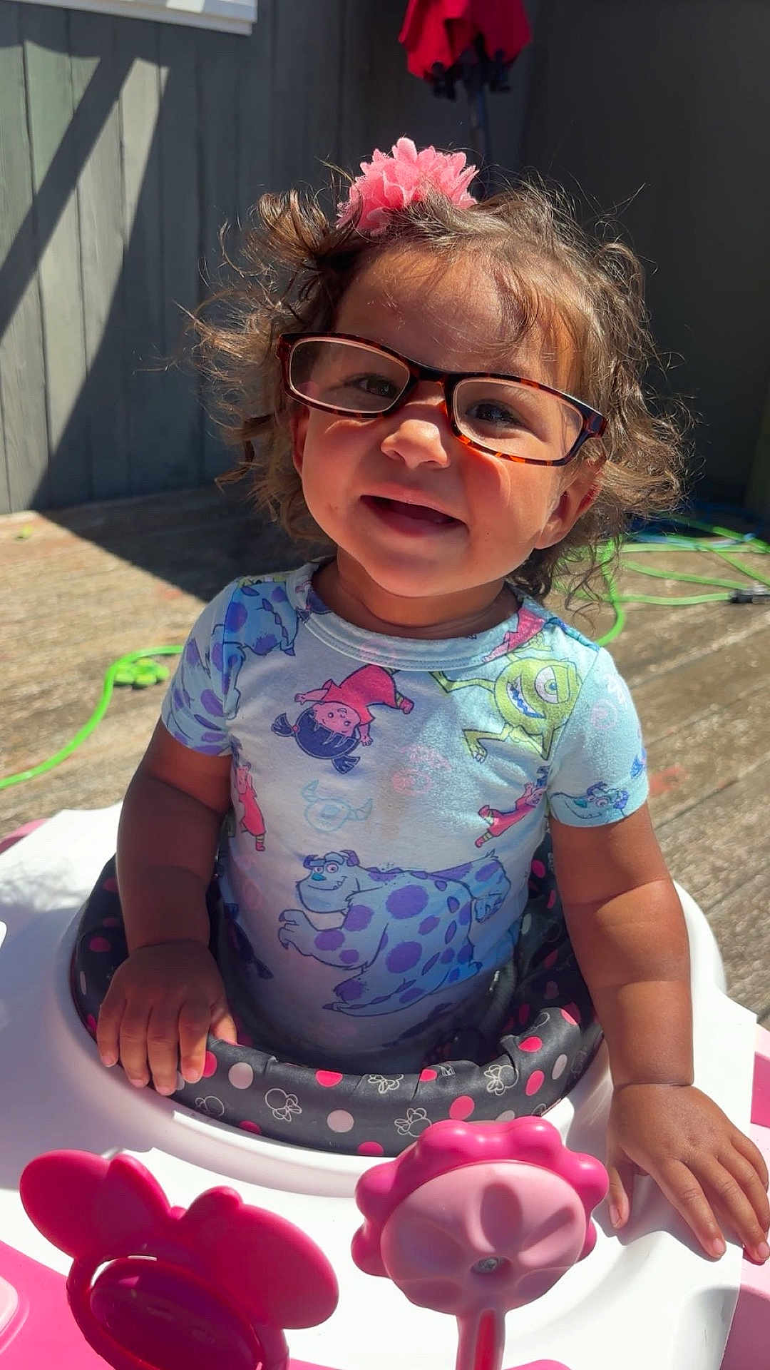 Kyelynn is registered to the contest to win money with this photo: toddler, child, glasses, pink, baby_walker, flower_toys, curly_hair, outdoor, sunlight, smiling, shirt, cartoon_print, hand, deck, happy, play, person, summer, cute, baby