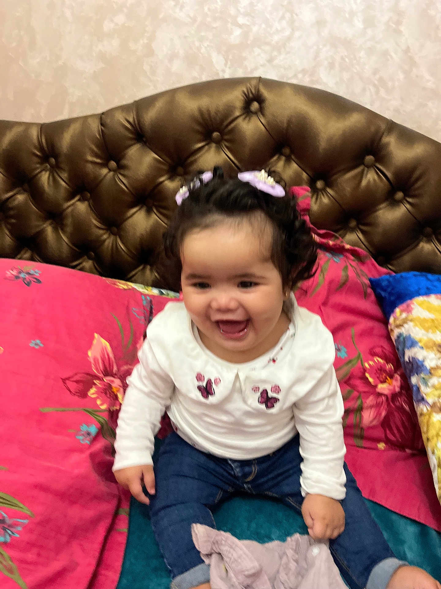 Emna participe au concours pour gagner de l'argent avec cette photo : toddler, child, smiling, curly_hair, hair_clips, white_shirt, jeans, bed, pillows, floral_pattern, headboard, indoor, happy, sitting, person, cute, young_child, furniture, home, playful