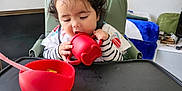 Emna participe au concours pour gagner de l'argent avec cette photo : toddler, child, high_chair, red_cup, red_bowl, spoon, curly_hair, two_buns, striped_clothing, feeding_time, indoor, living_room, toys, blanket, furniture, person, baby_feeding, cozy, home, cute