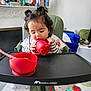 toddler, child, high_chair, red_cup, red_bowl, spoon, curly_hair, two_buns, striped_clothing, feeding_time, indoor, living_room, toys, blanket, furniture, person, baby_feeding, cozy, home, cute