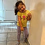 child, girl, smile, yellow_shirt, leggings, socks, wall, floor, tile_floor, cabinet, home, indoor, curly_hair, standing, leaning, happy, playful, person, casual_clothing, young_child