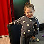 child, smiling, standing, heart_pattern, sweatshirt, leggings, indoor, chair, table, pacifier, remote_control, wood_floor, curtain, black_chair, happy, young_girl, casual_clothing, pony_tail, person, furniture