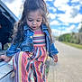 child, girl, curly_hair, denim_jacket, striped_outfit, sneakers, outdoor, daylight, clouds, sky, vehicle, road, sidewalk, casual_clothing, cute, portrait, young_child, walking, nature, playful