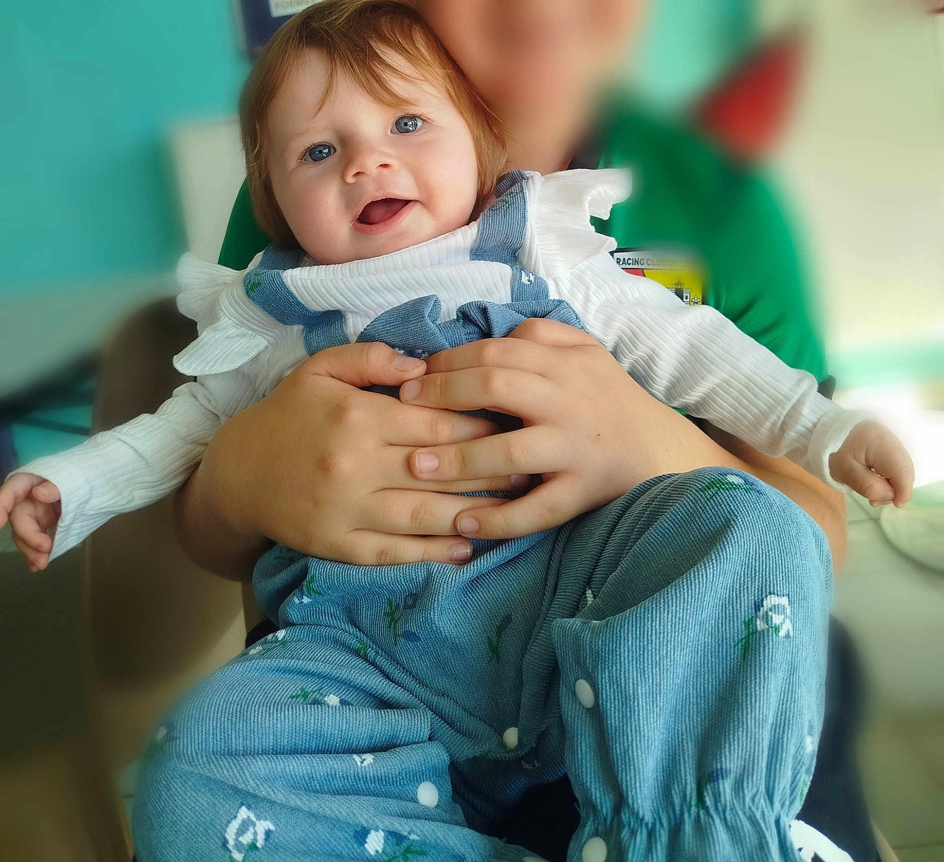 Joye participe au concours pour gagner de l'argent avec cette photo : baby, child, smiling, blue_eyes, overalls, white_top, hands, holding, indoor, person, happy, portrait, ruffled_sleeves, seated, blurred_face, blue_pants, floral_pattern, furniture, light, casual_clothing