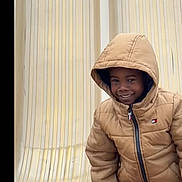 De Aire joined the competition — help win amazing prizes! child, smiling, portrait, hooded_jacket, winter_clothing, tan_coat, indoor, chair, plastic_chair, seated, standing, happy, face, person, candid, fashion, zipper, hood, black_background, photograph